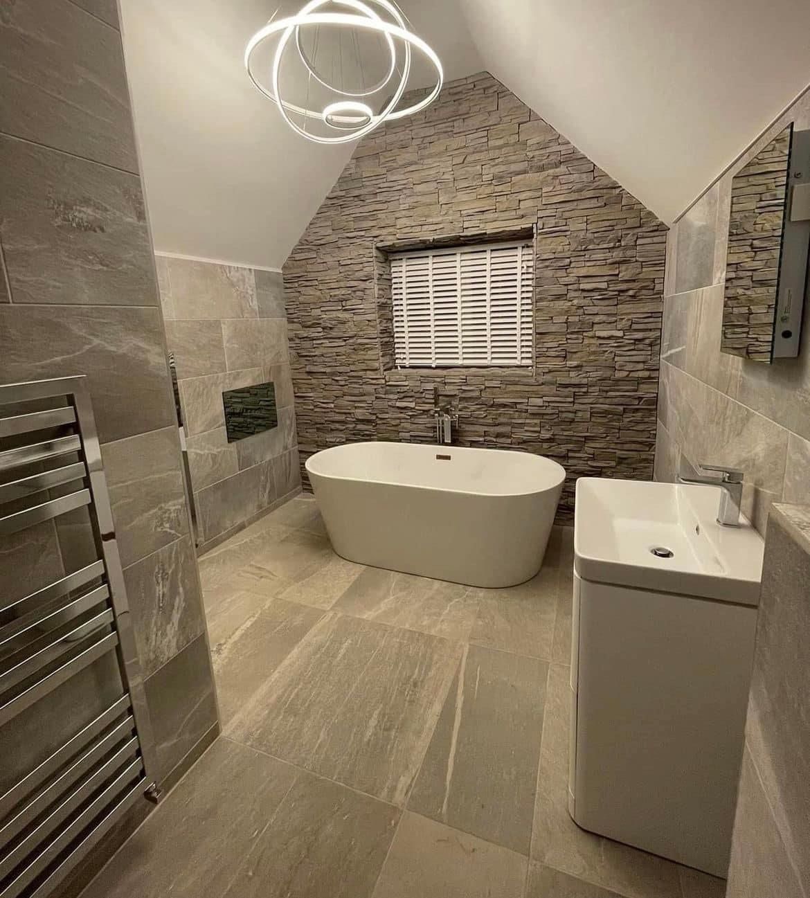A bathroom fitted by Gibson: freestanding bath under a sloped ceiling, stone feature wall and a ring light